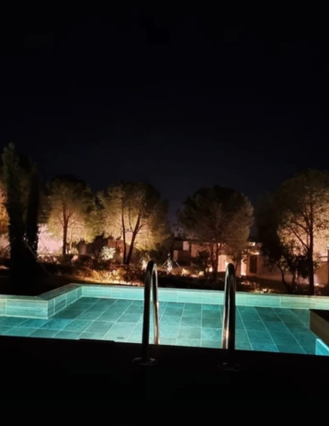 Gece aydınlatılmış turkuaz havuz ve arkada ışıklandırılmış çamlar