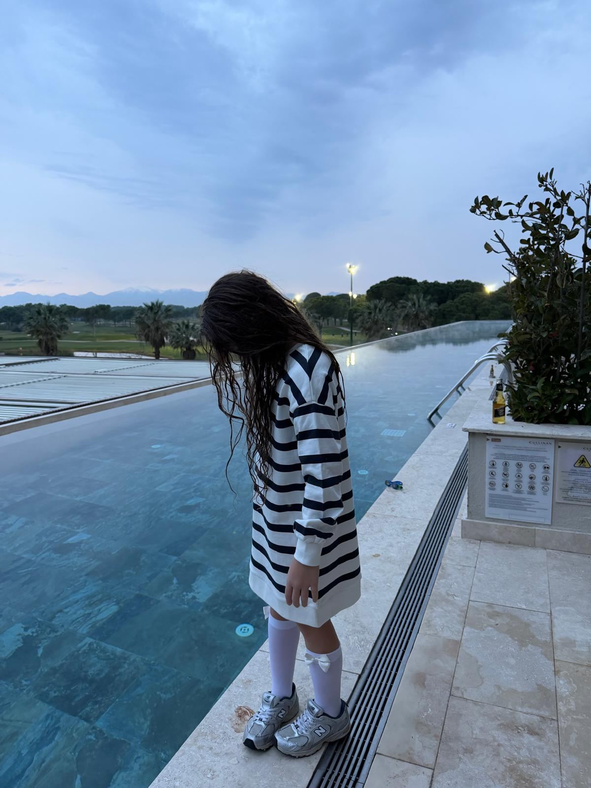 Havuz kenarında duran çizgili elbiseli bir çocuk; arkada golf sahası ve dağlar, havuz suyunda buhar