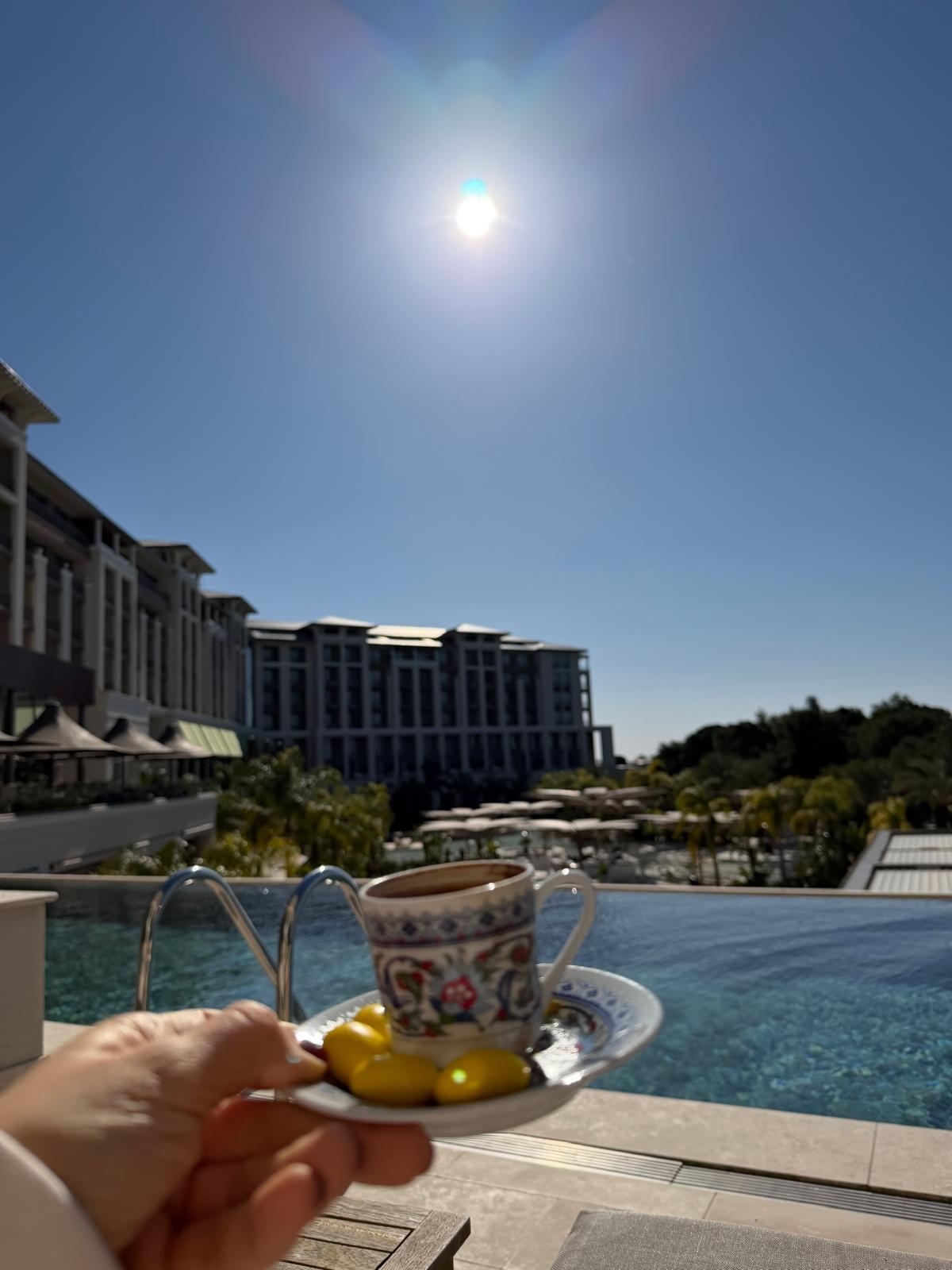 Sonsuzluk havuzunun kenarında elde tutulan desenli fincanda Türk kahvesi, arkada otel binaları