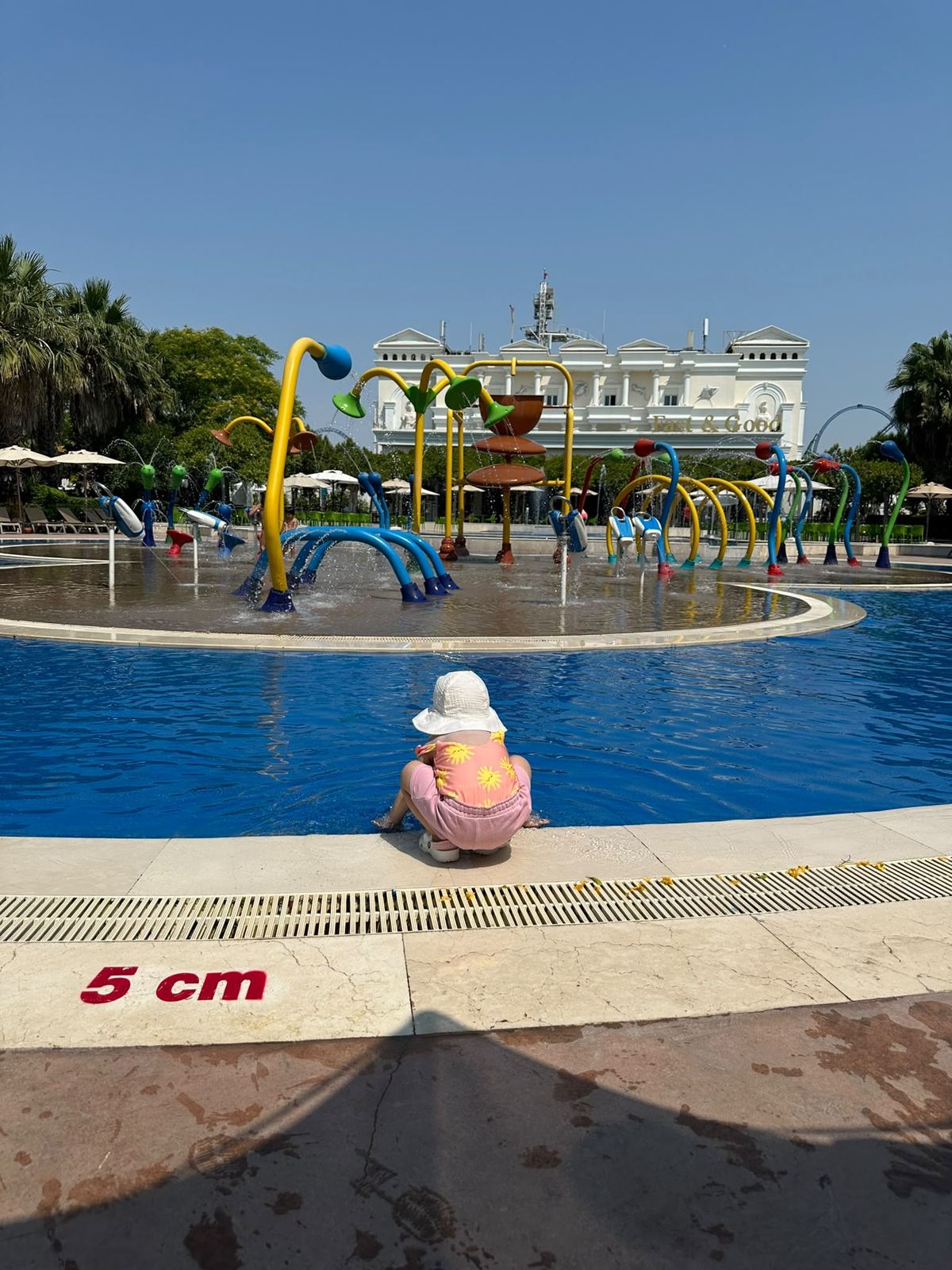 A shallow baby pool in front of the splash playground; a child in a sun hat crouches at its edge with their back to the camera, a 5 cm depth marker on the ground