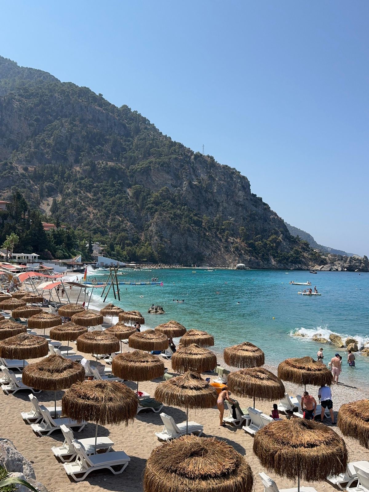 Hasır şemsiyeli çakıllı plaj, turkuaz koy ve arkada kayalık yamaç