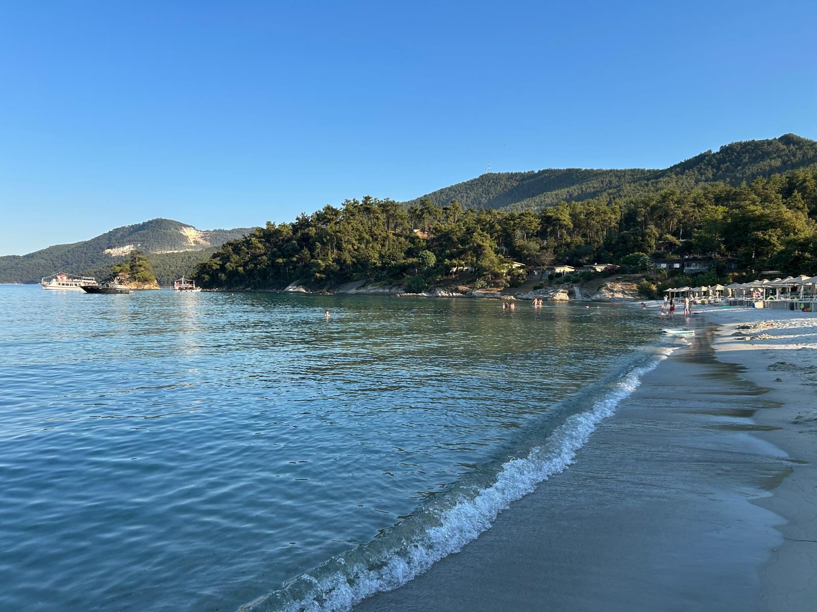 Kumlu kıyıya yumuşakça vuran berrak dalga, arkada çam kaplı tepeler ve tekneler