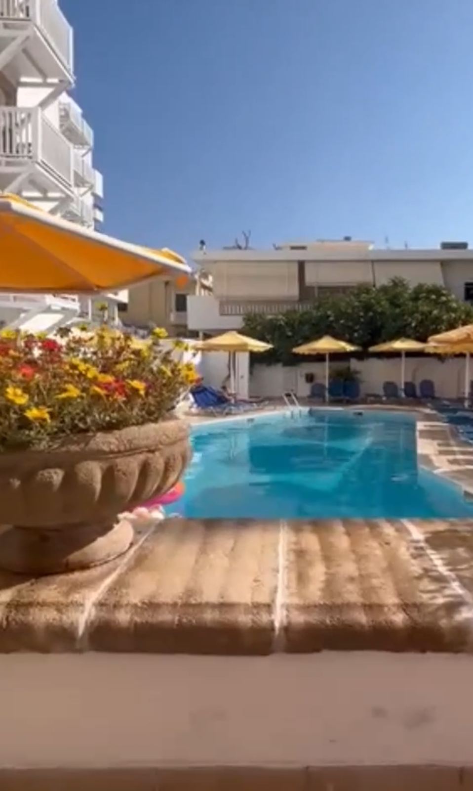 Çiçek saksılı terastan havuza bakış, sarı şemsiyeler ve otel binası