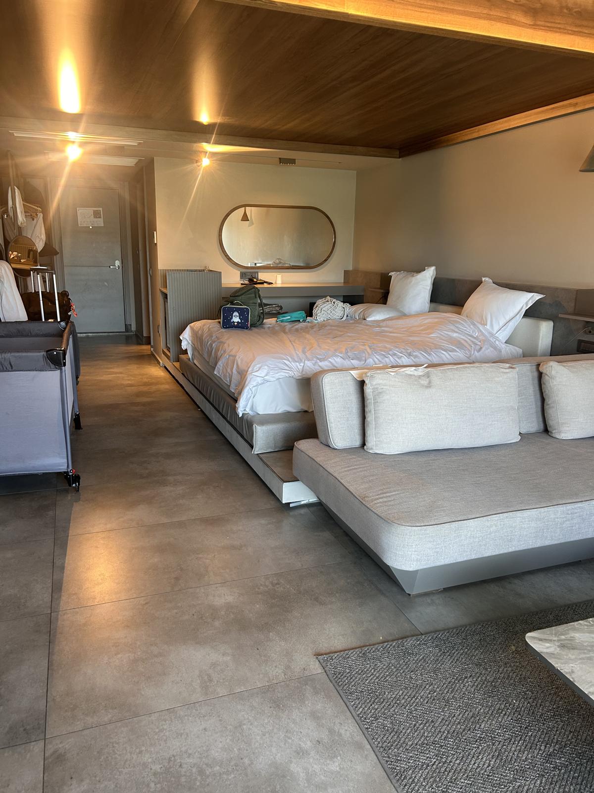 Otel odası; önde gri bebek seyahat yatağı, arkada yatak ve ayna