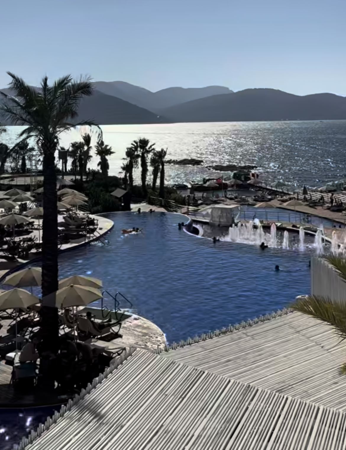 Palmiyeli geniş havuz, şemsiyeli teras ve arkada deniz ile dağ manzarası