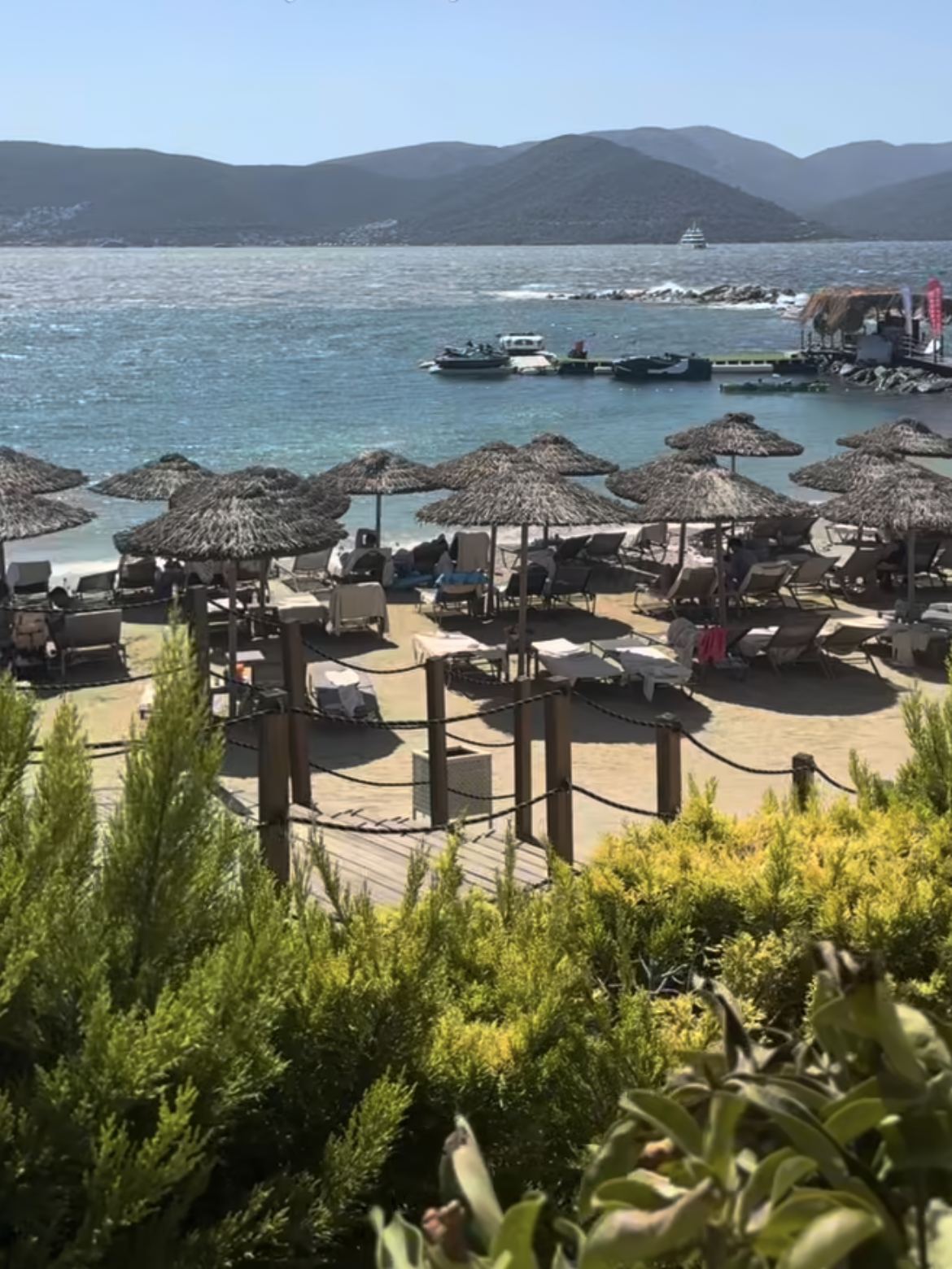 Hasır şemsiyeler, şezlonglar ve arkada deniz, iskele ve dağlar