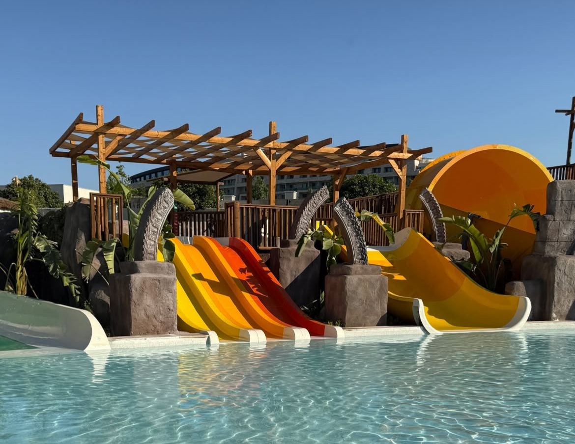 Sığ çocuk havuzuna inen turuncu-sarı mini kaydıraklar, arkada ahşap pergola